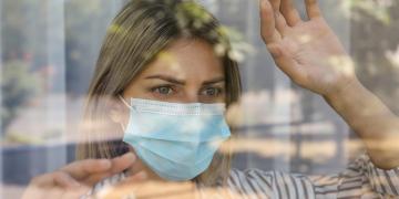Stressed woman in protective mask looking out of window, view through glass. Self-isolation during COVID-19 pandemic